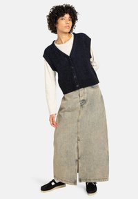 Marine gebreide mouwloze trui met korte mouwen over een crèmewit langemouwen shirt, gecombineerd met een lange denimrok en zwarte sneakers.