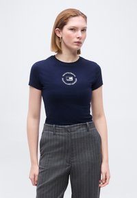 Γυναίκα με κοντά ξανθά μαλλιά, φοράει μια εφαρμοστή μπλε μπλούζα με κυκλικό λογότυπο και γκρι παντελόνι με ρίγες, στεκόμενη μπροστά σε ανοιχτό φόντο.