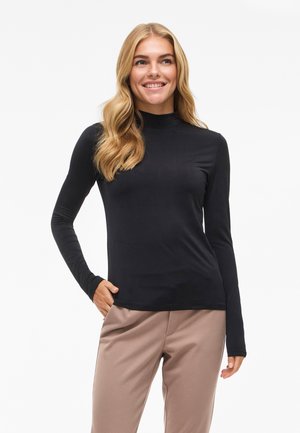 Maglietta nera a maniche lunghe con collo alto, dal design aderente, realizzata in un tessuto morbido, abbinata a pantaloni beige. Texture morbida e finitura senza cuciture.