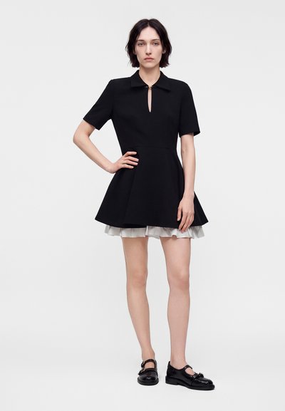 Femme en robe noire à manches courtes avec une jupe évasée et un ourlet à volants blancs, portant des chaussures Mary Jane noires et se tenant avec une main sur la hanche.