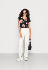 Top preto de mangas curtas com padrão floral combinado com calças largas em creme, ténis brancos e uma pequena bolsa preta. O modelo tem cabelo encaracolado e óculos de sol.