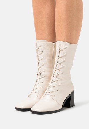 Bílé boty kotníkové z umělé krokodýlí kůže s šněrováním na přední straně, špičatou špičkou a blokovým podpatkem. Obsahují boční zip pro snadné obouvání.