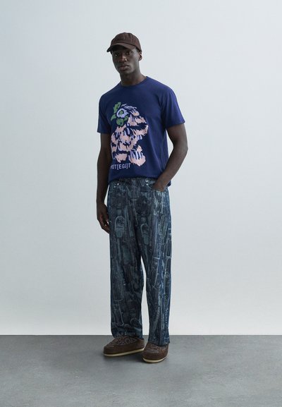 Μπλε ναυτικό γραφικό μπλουζάκι με λουλουδένιο σχέδιο, συνδυασμένο με φαρδιά μπλε παντελόνια με μοτίβο και καφέ παπούτσια τύπου slip-on. Καφέ καπέλο ολοκληρώνει την εμφάνιση.