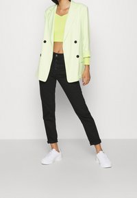 Limonkowa, podwójnie zapinana marynarka, czarne dopasowane dżinsy i białe sneakersy. Marynarka ma czarne guziki i podwinięte rękawy.