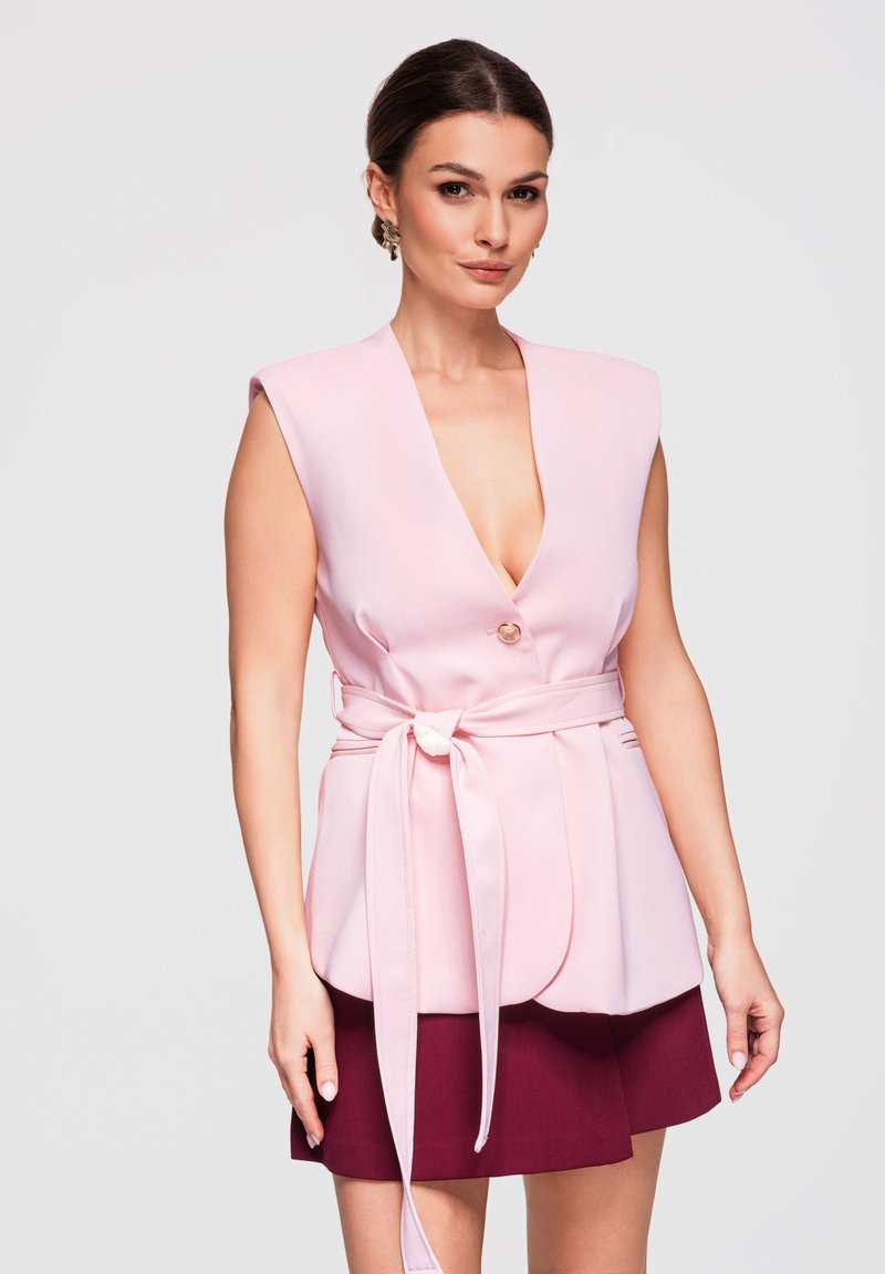 Femme portant un blazer sans manches rose clair ceinturé avec un col en V profond, sur une jupe rouge foncé, debout devant un fond clair uni.