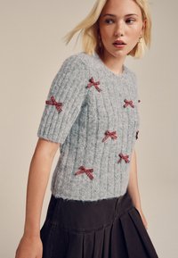 Pull en maille côtelée gris clair avec manches courtes et nœuds en tissu écossais rouge décorant le devant. Texture douce, coupe ajustée.