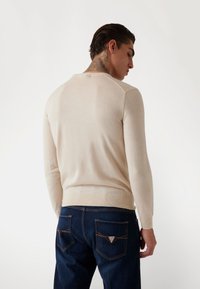 Helle beige Langarmbluse aus feinem Strickstoff, mit gerippten Bündchen und Saum. Kombiniert mit dunkelblauen Jeans, die eine dreieckige Gesäßtasche haben.