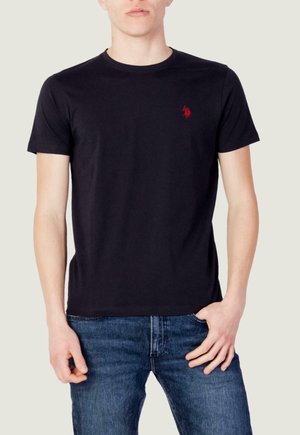 Homme portant un t-shirt noir uni à manches courtes et col rond avec un petit logo rouge sur la poitrine gauche, associé à un jean bleu, debout avec une main dans la poche.