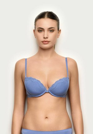 Kobieta z włosami zaczesanymi do tyłu, ubrana w jasnoniebieski biustonosz z koronkowym wykończeniem i dopasowane majtki, zwrócona przodem na neutralnym tle.