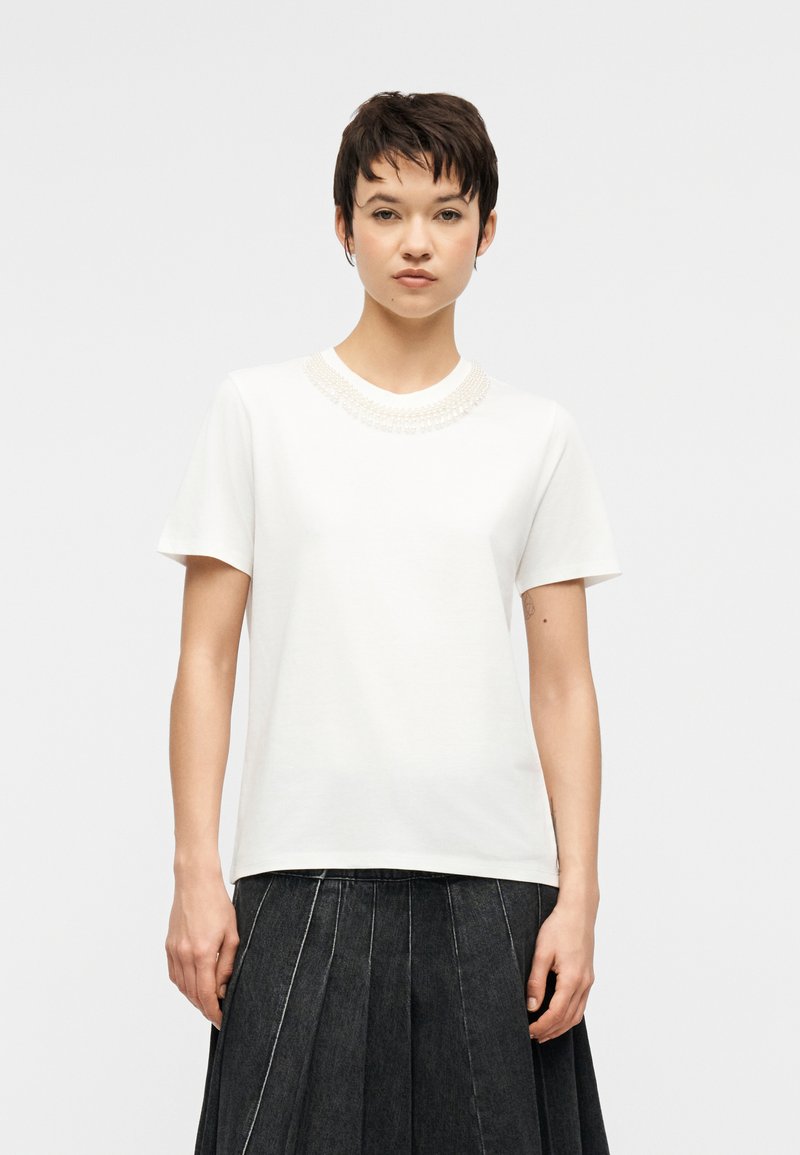 Νεαρό άτομο με κοντά σκούρα μαλλιά φορούσε λευκό κοντομάνικο τοπ με κέντημα στο λαιμό και μαύρη πλισέ φούστα, στέκεται μπροστά σε απλό φόντο.