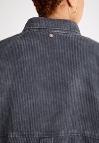 Zadný pohľad na šedú kordurovú košeľu s gombíkom na golieri, s vertikálnou rebrovou textúrou a mierne rozšíreným spodným okrajom.