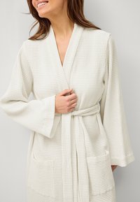 Robe blanc avec une texture gaufrée, col châle, manches longues et ceinture à la taille. Dotée de deux poches avant et d'un tissu doux et léger.