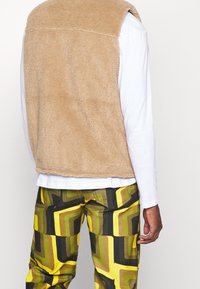 Tan fleeceväst med mjuk textur, i kombination med mönstrade byxor med geometriska mönster i gult, brunt och svart.