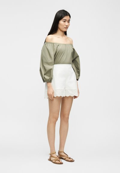 Γυναίκα με μακριά σκουρόχρωμα μαλλιά, που φοράει πράσινο ελιάς μπλούζα με ανοικτούς ώμους, λευκά σορτς με δαντέλα και καφέ σανδάλια με λουριά, σε πλευρική στάση.