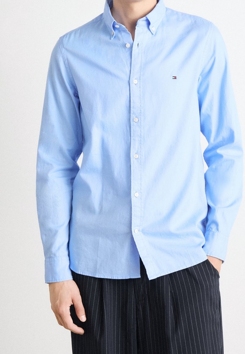 Personne portant une chemise bleu clair à boutons avec un petit logo sur la poitrine et un pantalon noir à fines rayures, debout avec une main dans une poche.