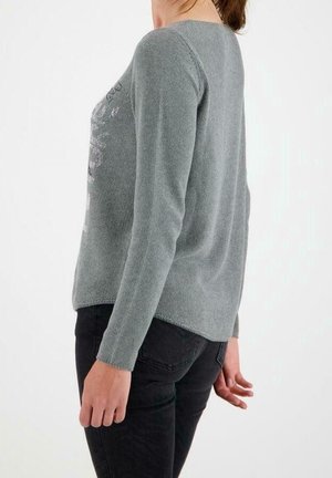 Donna che indossa un maglione grigio chiaro a maniche lunghe e pantaloni neri, in piedi di profilo su uno sfondo bianco semplice.