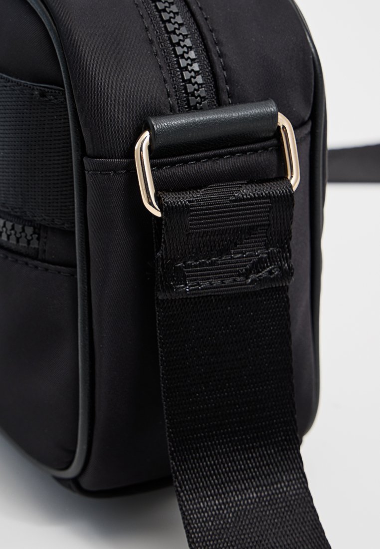 Bolso de tela negra con cierre de cremallera, correa texturizada y acentos metálicos. Presenta una forma rectangular elegante y una superficie lisa.