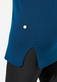 Maglione lavorato a maglia color teal con orlo laterale e bordi scallop, con un piccolo dettaglio di bottone dorato sul lato sinistro. Abbinato a pantaloni neri.