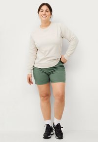 Beige langærmet skjorte med præget tekst, parret med grønne shorts. Modellen bærer sorte sportssko og hvide sokker. Diskret, afslappet design.