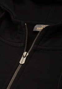Felpa nera con zip dotata di una cerniera dorata e un'etichetta taggata, realizzata in tessuto morbido e con cappuccio. Dettagli includono un tirazip personalizzato.