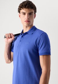 Plava polo majica od teksturiranog materijala, sa tri dugmeta na prednjem dijelu i izvezenim logom. Ovratnik ima kontrastne pruge.