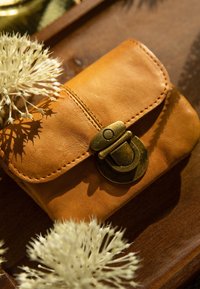 Brązowy skórzany portfel z zaokrąglonym klapkiem i mosiężnym zapięciem, z widocznymi szwami i gładką teksturą, leżący na drewnianej powierzchni z kwiatami.