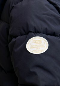 Marineblå pufferjakke med quiltet struktur, prydet med et rundt hvidt stofmærkat med beige tekst og en syet kant.