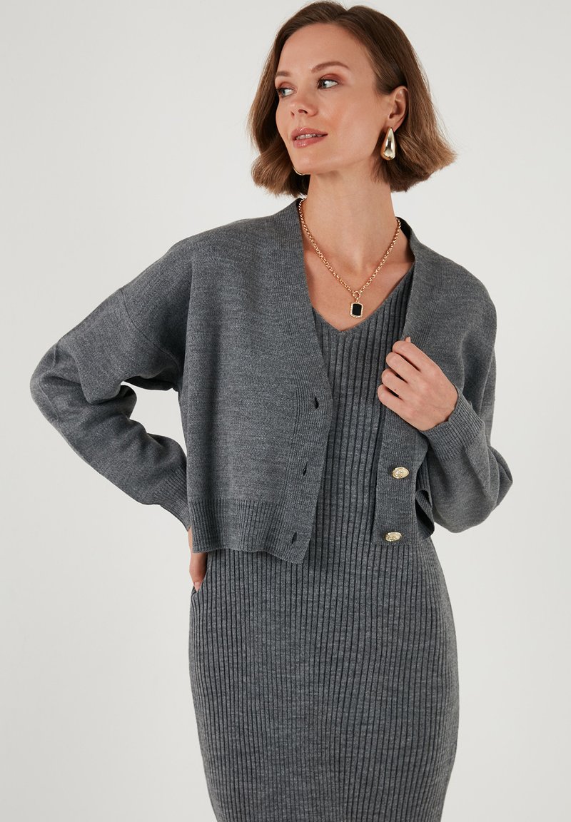 Cárdigan gris de punto con mangas largas y botones, llevado sobre un vestido gris de canalé. Textura suave con un corte relajado y detalles de diseño sutiles.