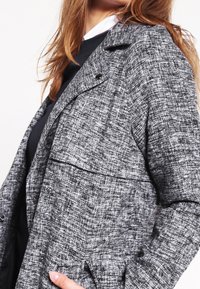 Giacca grigia dal tessuto testurizzato con un motivo in bianco e nero, caratterizzata da un colletto a scollo a V, chiusura con bottoni e tasche laterali.