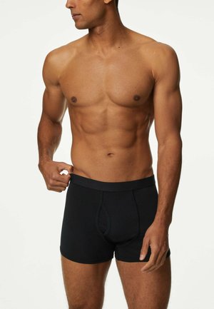Schwarze Boxershorts aus Baumwolle mit snug fit. Der Bund ist elastisch und hat ein schlichtes Design. Das Modell posiert mit einer Hand auf dem Bund.