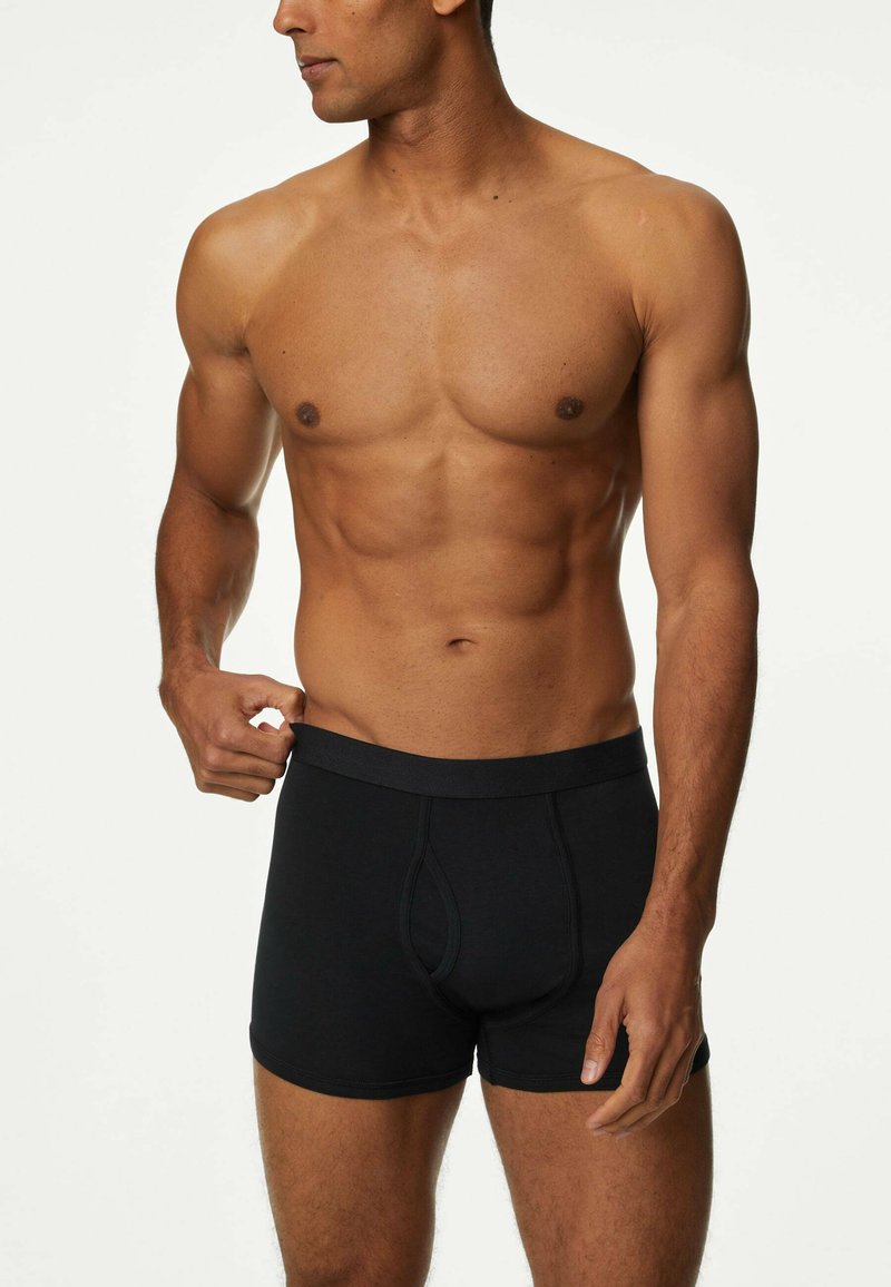 Czarne bokserki wykonane z bawełny, o dopasowanym kroju. Pasy elastyczny z prostym wzorem. Model pozuje z jedną ręką na pasie.