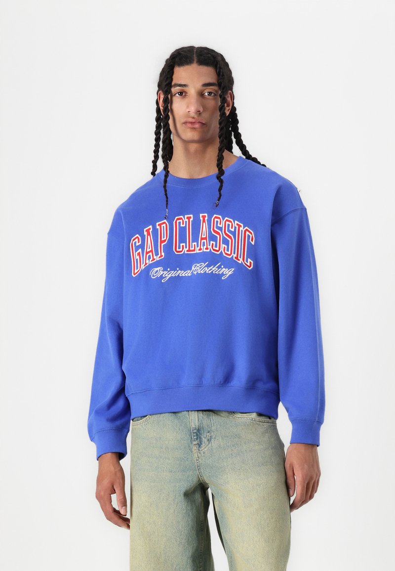Sudadera azul con el gráfico "GAP CLASSIC" en letras rojas y blancas, con un corte relajado y diseño de cuello redondo. Combinada con unos vaqueros de denim claros.