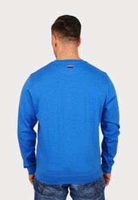 Homme portant un sweat-shirt bleu vif à manches longues et un jean bleu, debout dos à la caméra sur un fond clair uni.