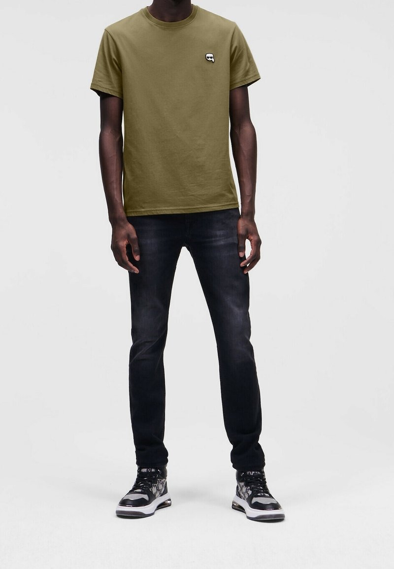 Olivgrünes Baumwoll-T-Shirt mit kurzen Ärmeln und kleinem Logo auf der Brust, kombiniert mit dunkler Denim-Jeans und schwarzen Turnschuhen.