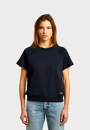 Vrouw met stijlbruin haar, die een zwart sweatshirt met korte mouwen en lichtblauwe spijkerbroek draagt, staand tegen een effen achtergrond.