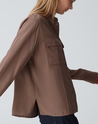 Bruine langemouwenhemd met een knoopsluiting en twee borstzakken. Gladde textuur, licht oversized ontwerp, zijsplitten aan de zoom.