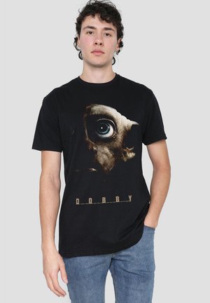 Mann trägt schwarzes T-Shirt mit großer Grafik eines Kreaturenauges und Gesichts sowie dem Wort "DOBBY" darunter, kombiniert mit blauen Jeans.