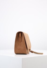 Bolso de ante color marrón claro con un diseño estructurado, que presenta un cierre con solapa y un acento de borlas. Textura suave y líneas limpias.