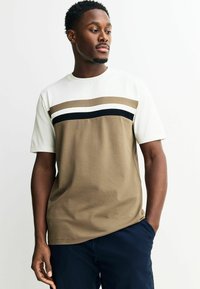 Man met kort krullend haar, gekleed in een beige-wit kleurgeblokte T-shirt met zwarte streep, marineblauwe broek, kijkt opzij met één hand in zijn zak.