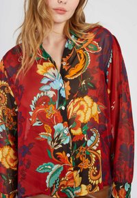 Durchsichtige rote Bluse mit floralem Muster in verschiedenen Farben, mit grünen Akzenten und weißen Knöpfen, im lässigen Schnitt und mit langen Ärmeln.