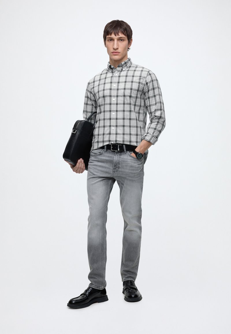 Unge mand iført en grå ternet skjorte, grå jeans, sorte sko og holder en sort lædertaske mod en hvid baggrund.