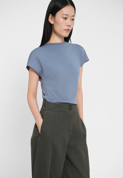 Μπλε τοπ με κοντά μανίκια και ψηλό λαιμό, συνδυασμένο με ψηλόμεσες σκούρες πράσινες παντελόνες που διαθέτουν διπλό κλείσιμο με κουμπί και πλαϊνές τσέπες.