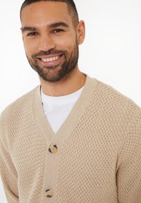 Beige gestrickte Cardigan mit strukturiertem Muster, V-Ausschnitt und drei Knöpfen. Über einem weißen T-Shirt getragen, mit glatter Verarbeitung.