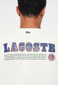 Balta kokvilnas T-kreklu ar krāsaino "LACOSTE" tekstu, izšūtu zaļu krokodilu logo un izdrukātu tekstu ar zilu tenisa mantojuma emblemu.