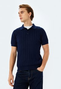 Marinblå stickad polo med kort ärm, med vertikal ribbad struktur och klassisk krage, tillsammans med mörka denimjeans.