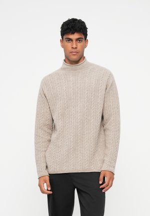 Pull en maille beige avec un col haut et des manches longues, présentant un design texturé, porté avec un pantalon sombre.