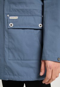 Blå jacka med en slät textur, med en sidficka, tryckknappar och en etikett. Synlig sömnad längs sömmarna.