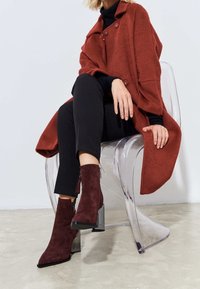 Mujer sentada en una silla transparente, vestida con un abrigo color óxido, pantalones negros, top negro y botas de tacón en ante marrón con cremalleras laterales.