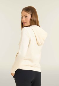 Donna con capelli castano chiaro che indossa un hoodie color crema e pantaloni neri, rivolta leggermente di lato su uno sfondo chiaro e semplice.