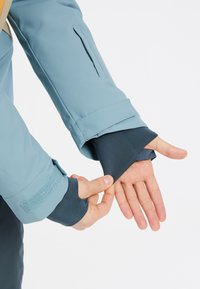 Šviesiai mėlynas vandeniui atsparus striukės rankovas su struktūruotu tamsiai mėlynu manžetu. Yra velcro dirželis ir paslėpta kišenė ant rankos.